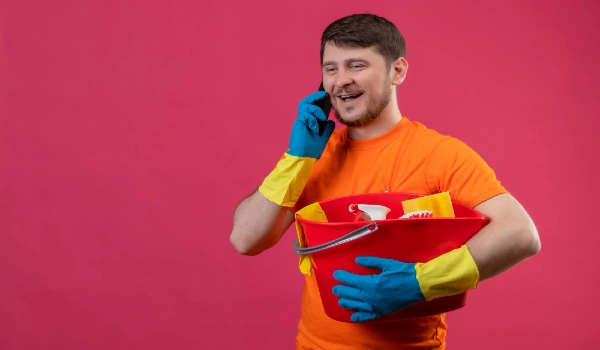Young man wearing orange t-shirt and rubber gloves holding bucket with cleaning tools