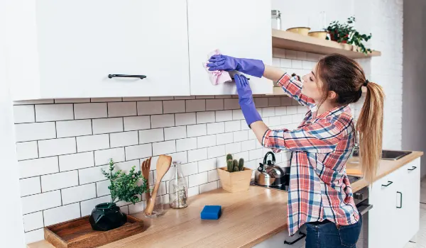 Woman in gloves cleaning cabinet with rag at home kitchen.