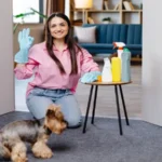 Young woman sitting near the table with cleaning products at home and her little dog is running next to her