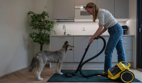Young woman vacuuming carpet with her dog at kitchen