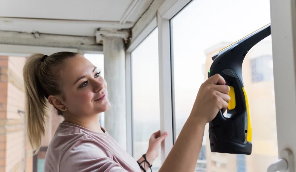 Beautiful young housewife in working clothes washing windows by professional tools at home